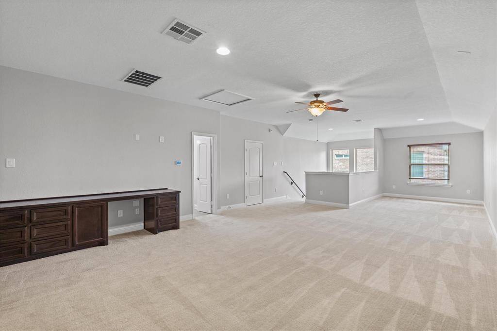 Spacious, bright room with neutral colors, featuring plush carpet, a ceiling fan, built-in desk, and multiple windows providing natural light. Ideal for a family room or home office.