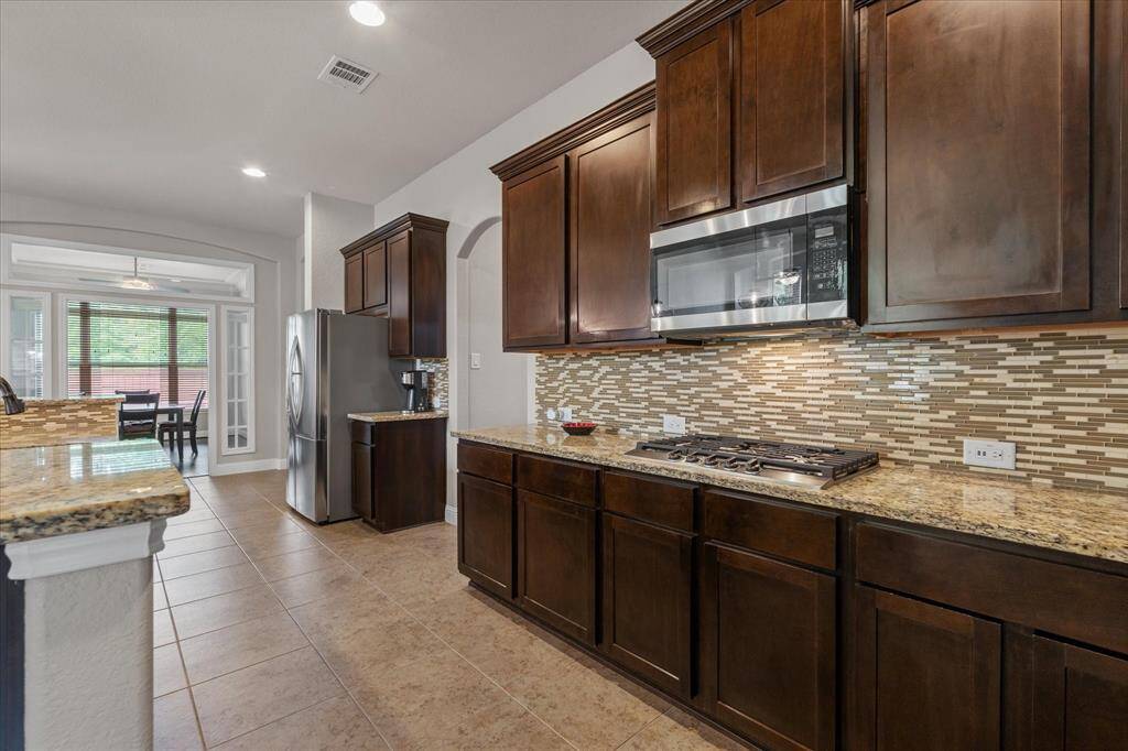This kitchen features dark wood cabinetry, granite countertops, and a modern tile backsplash. It includes a stainless steel microwave and gas stove. The space is open with a view into a dining area framed by large windows, allowing natural light to fill the room.