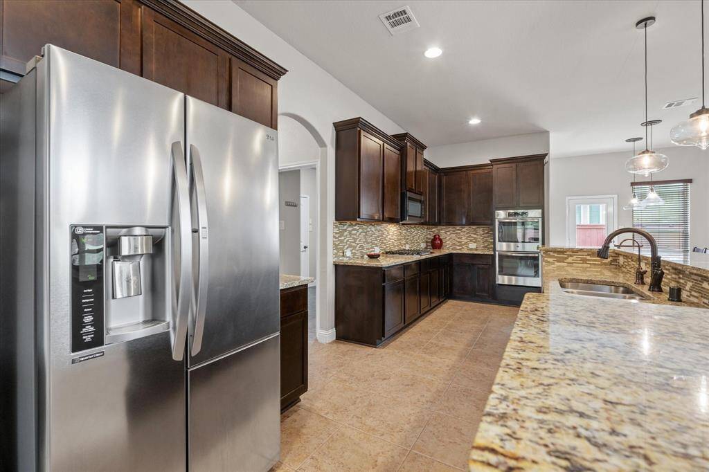Modern kitchen featuring stainless steel appliances, dark wood cabinetry, granite countertops, and stylish pendant lighting. The open layout and tiled flooring add a spacious and elegant touch.