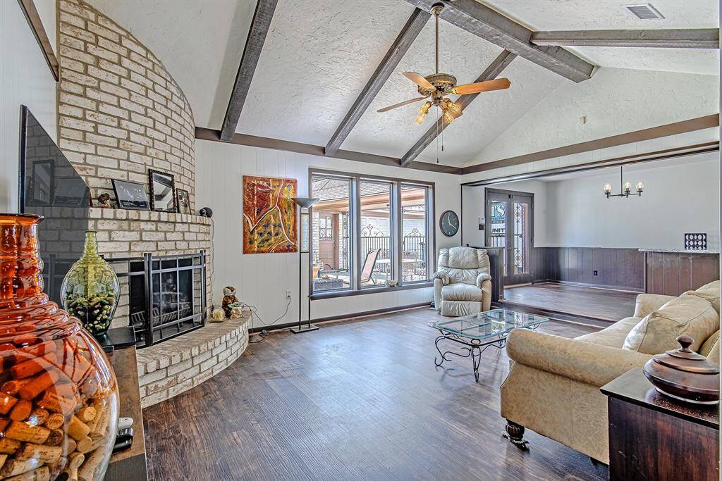 Spacious living room with vaulted ceilings, exposed beams, and a stunning brick fireplace, flowing seamlessly into the dining room, perfect for entertaining or relaxing.