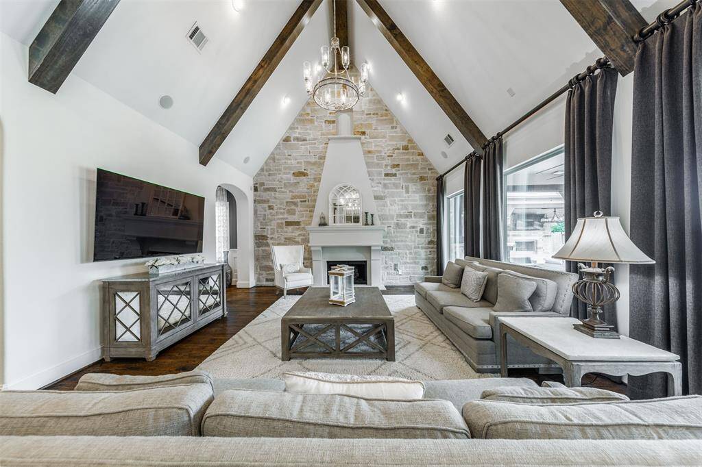 The impressive family room sits beneath a stunning cathedral ceiling with rustic wood beams, creating a warm and inviting atmosphere. An elegant fireplace set against a striking stone display wall serves as the focal point, while large windows flood the room with natural light and offer beautiful views of the outdoor oasis.