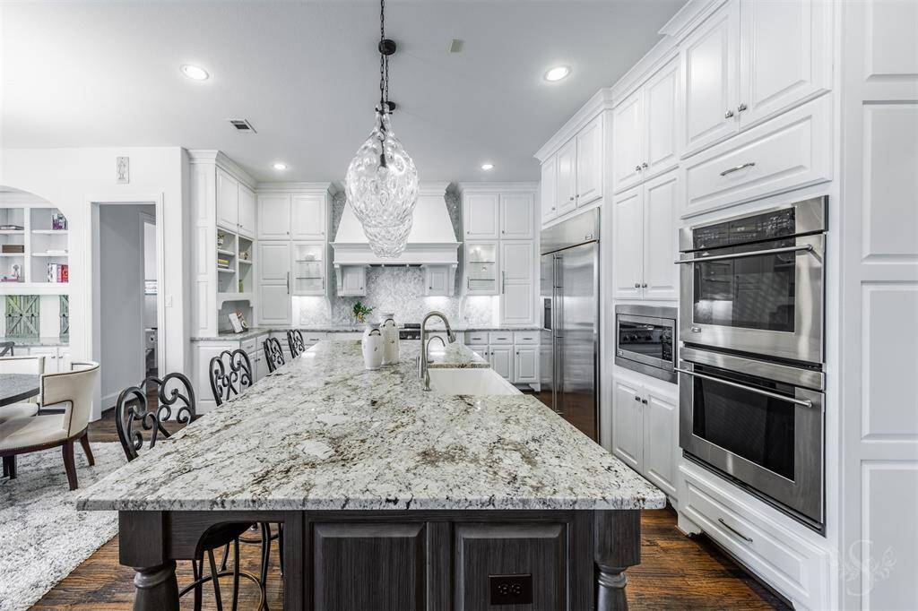 This modern, upscale kitchen is as functional as it is stunning, anchored by a massive granite island that doubles as a breakfast bar, perfect for casual dining or entertaining. Featuring custom pendant lighting, an abundance of crisp white cabinetry, including elegant glass-front accents, high-end stainless steel appliances including double ovens, and a thoughtful layout make this space a chef’s dream and a true centerpiece of the home.