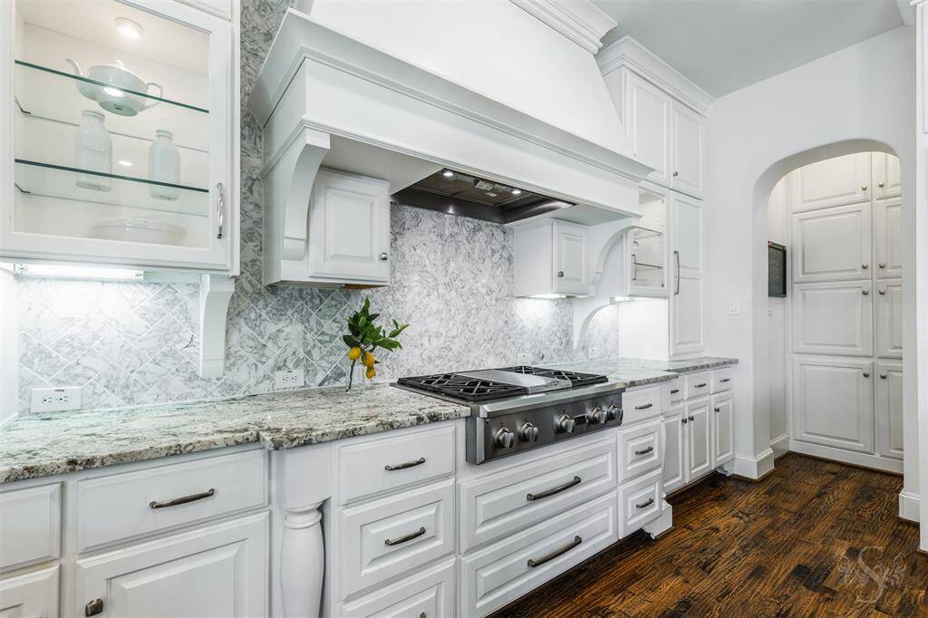 The gas cooktop is framed by elegant granite countertops and a stunning tile backsplash, creating a stylish cooking area. Glass-faced white cabinetry and warm wood flooring complete the look, giving the kitchen a clean, inviting feel.