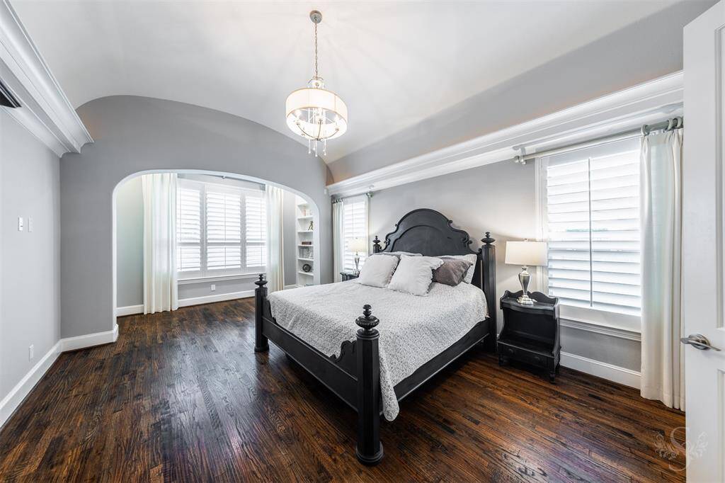 A serene primary bedroom boasts beautiful wood flooring and an elegant arched ceiling with crown molding, adding a touch of sophistication. Plantation shutters let in just the right amount of light, while a cozy sitting room with built-ins offers a perfect space to relax or unwind.