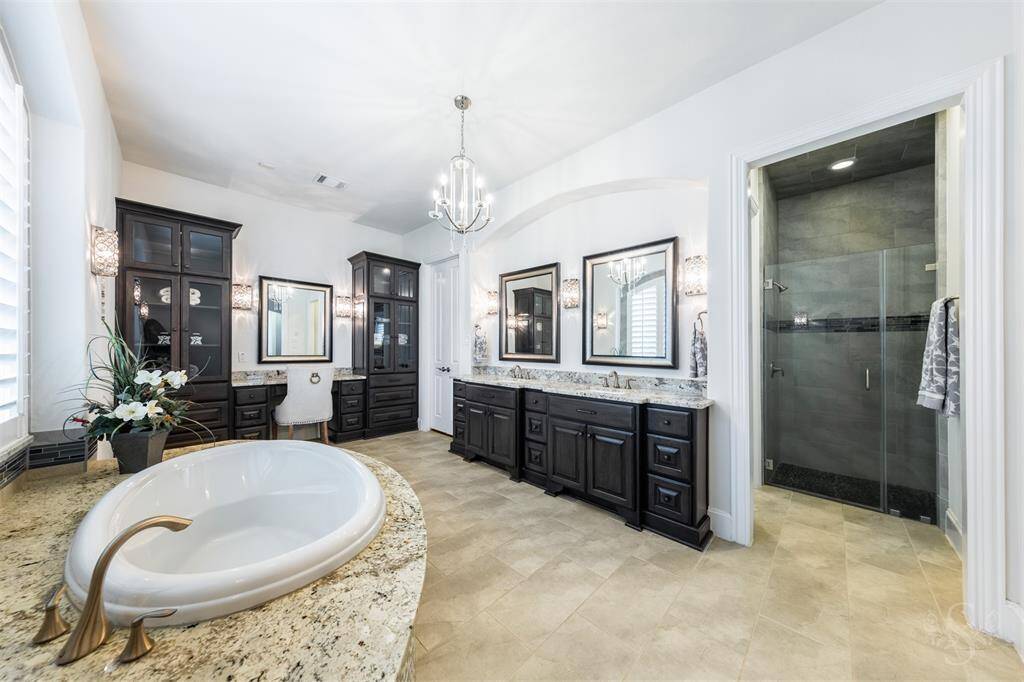 A luxurious primary en-suite bath feels like a spa, featuring a beautiful soaking tub surrounded by granite and decorative tile, perfect for unwinding. The extended double sink vanity with granite countertops and framed mirrors, chandelier, enclosed shower along with a make-up counter and built-in cabinets with glass fronts, adds both style and convenience to the space.