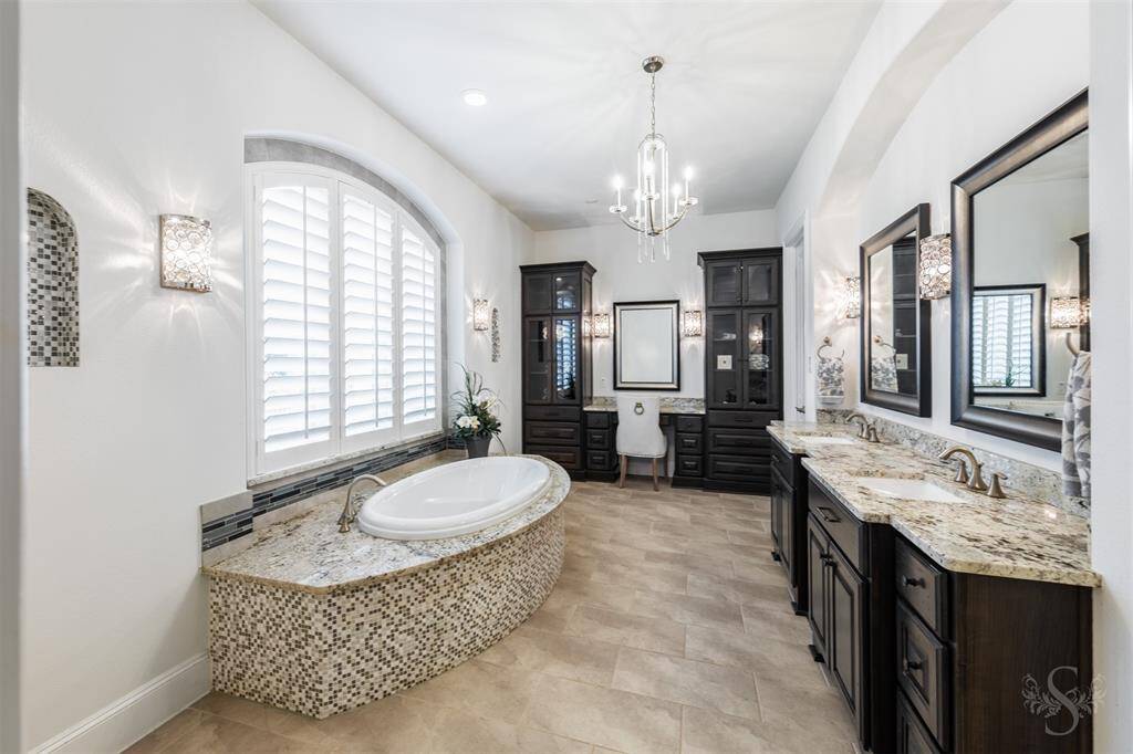 A luxurious primary en-suite bath feels like a spa, featuring a beautiful soaking tub surrounded by granite and decorative tile, perfect for unwinding. The extended double sink vanity with granite countertops and framed mirrors, chandelier and enclosed shower along with a make-up counter and built-in cabinets with glass fronts, adds both style and convenience to the space.