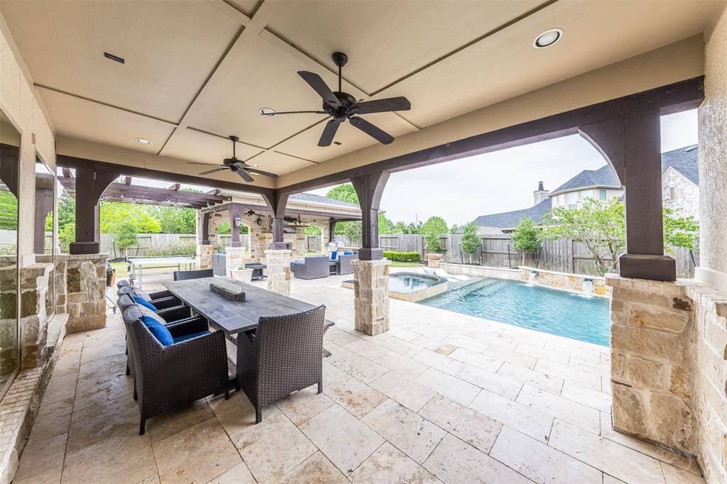 The inviting outdoor living area features a covered patio with recessed lighting and ceiling fans, perfect for enjoying the outdoors in comfort. Stone posts frame the space, which overlooks a sparkling pool and spa, creating a relaxing retreat for both lounging and entertaining.