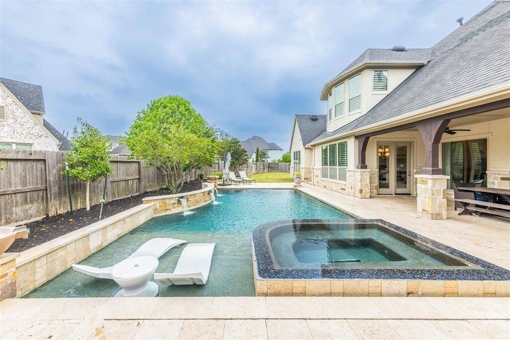 This stunning home pool features a relaxing tanning deck, a soothing spa, and a beautiful waterfall, creating a perfect oasis for relaxation. Lush landscaping behind the pool adds a touch of nature, enhancing the tranquil vibe of the outdoor space.