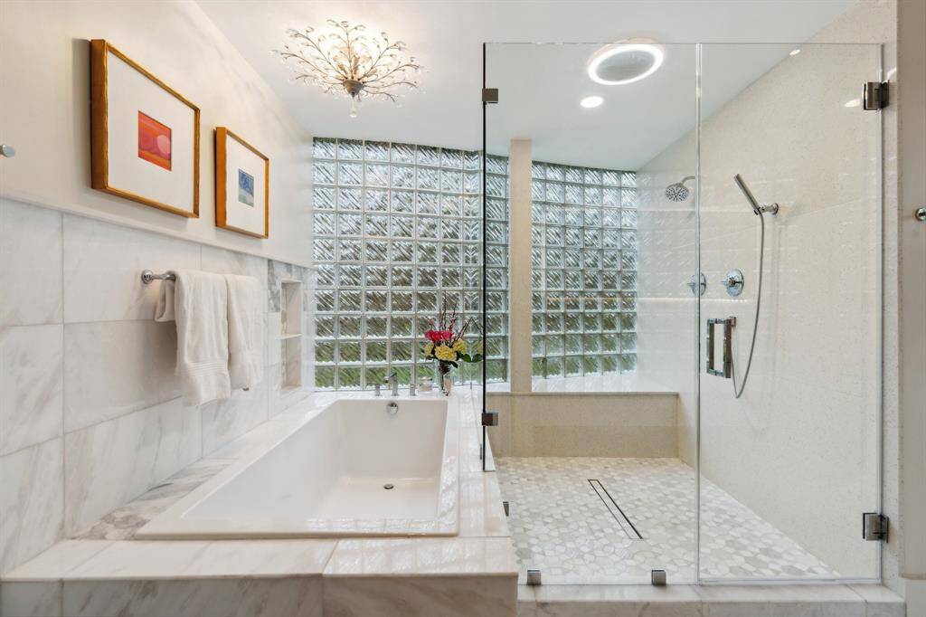 The oversized primary  shower, featuring a bench, handheld spray, and decorative tile floor, is complemented by a separate soaking tub with marble surround. The water closet is separate for added privacy, and marble tile floors complete the look. Not shown is the massive walk-in closet which includes an island with storage, cabinets, linen storage, shoe shelves, and a decorative light fixture.