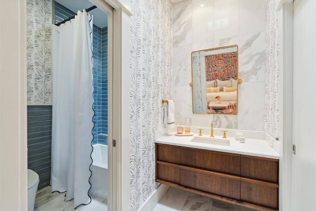 The ensuite bathroom for Secondary Bedroom #1 includes a floating vanity with a brass plumbing fixture and quartz countertop. Decorative wallpaper creates a chic ambiance. Separate space with a toilet and tub/shower combination featuring a stacked tile wall. Marble tile floors and a hidden medicine cabinet add both functionality and style.  A pocket door to the right opens to a walk-in closet.