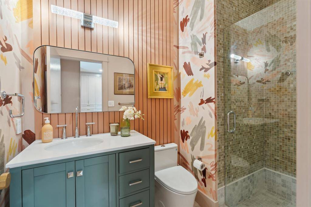 The ensuite bathroom for Bedroom #4 features elegant marble floors, a vanity with storage, and a decorative mirror. Schumacher wallpaper and a paneled accent wall add texture and warmth, while a decorative vanity light enhances the space. The walk-in shower, with its glass door and tiled surround, offers a clean and modern finish.