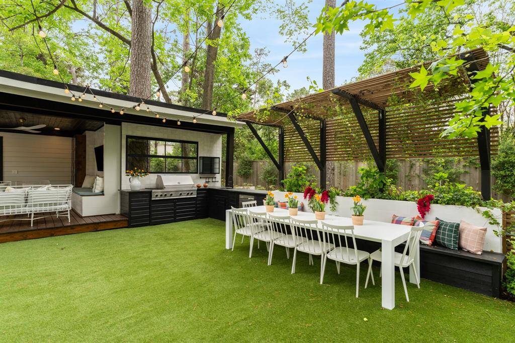 The outdoor kitchen is equipped with a stainless-steel grill, sink, mini-fridge, burner, storage, and a leathered granite countertop. A charming trellis hangs over the built-in banquette and dining area, while twinkling lights create an inviting atmosphere.