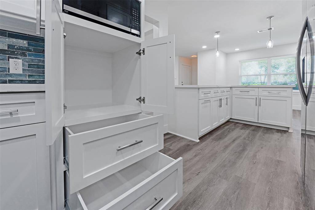 Modern kitchen with ample white cabinetry, sleek metal handles, and a built-in microwave. Features a stylish tile backsplash, wood-look flooring, and recessed lighting. Large window provides natural light, enhancing the spacious feel.