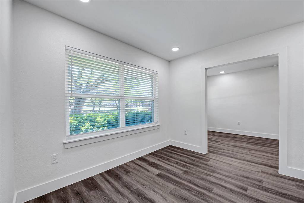 This photo shows a bright room with large windows featuring blinds, offering ample natural light. The space has a modern feel with light-colored walls and wood-style flooring. An open doorway leads to an adjacent room, enhancing the open layout.