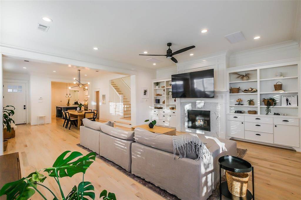 This photo showcases a bright, open-concept living and dining area with light wood floors and modern finishes. The living room features a cozy fireplace, built-in shelving, and a large TV. A stylish dining area and staircase are visible, enhancing the spacious feel.