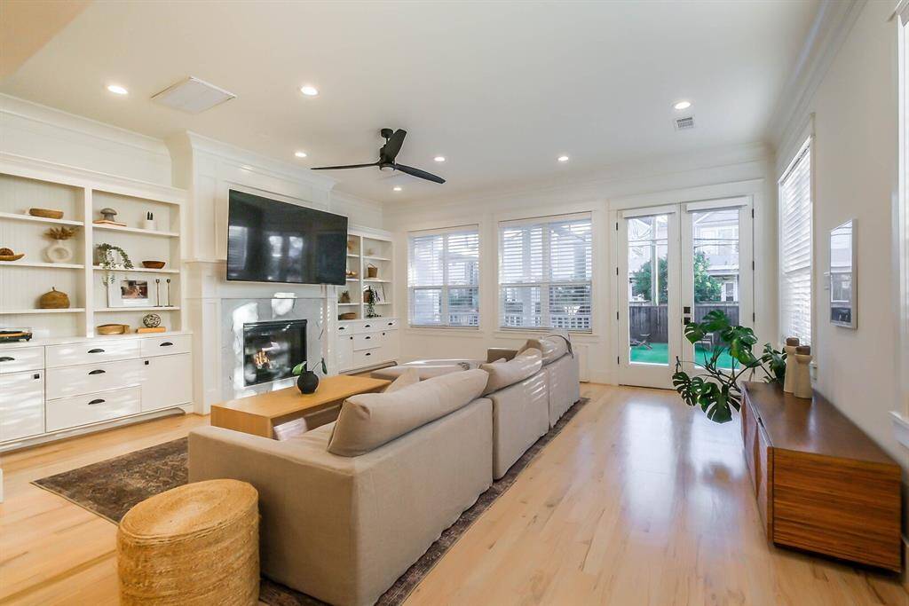 This inviting living room features a cozy fireplace, built-in shelving, and a mounted TV. Ample natural light streams in through large windows and French doors, leading to a patio. The space includes a ceiling fan for added comfort.