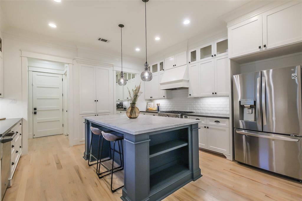 This kitchen features a contemporary design with white cabinetry, stainless steel appliances, and a central island with seating. Pendant lighting and light wood flooring add to the bright, modern feel.