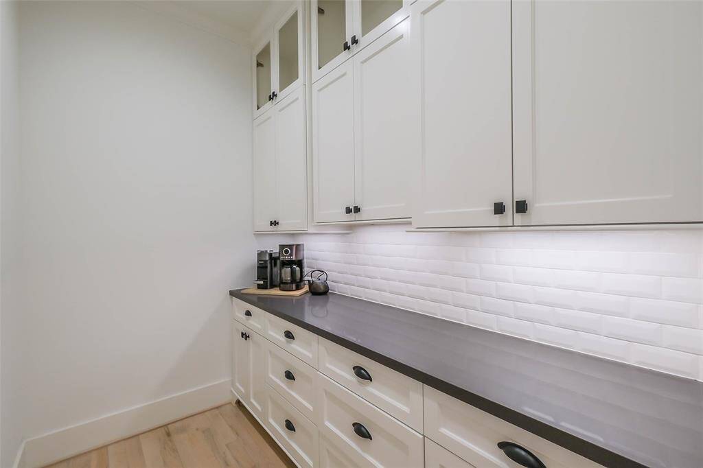 This photo showcases a sleek Butler's pantry or utility space with ample white cabinetry, black hardware, a modern countertop, and a white subway tile backsplash. Bright under-cabinet lighting enhances functionality.