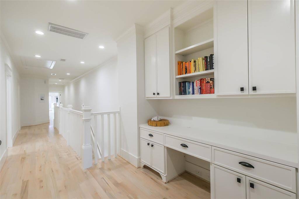This hallway features light wood flooring and white walls, leading to a built-in desk with upper and lower cabinets, ideal for a home office or study area. The space is bright and airy, with recessed lighting and a railing along the staircase.