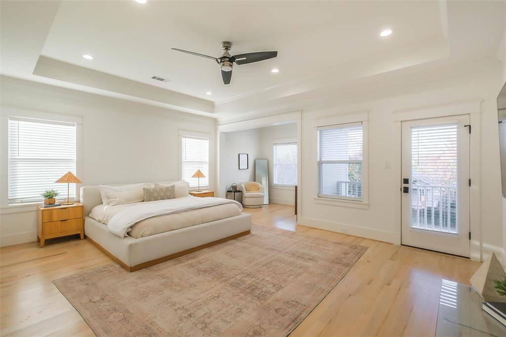 This spacious Primary bedroom suite features a modern design with light wood flooring and neutral walls. It includes ample natural light from multiple windows, and a cozy sitting area. The room also has a ceiling fan and access to a private balcony.