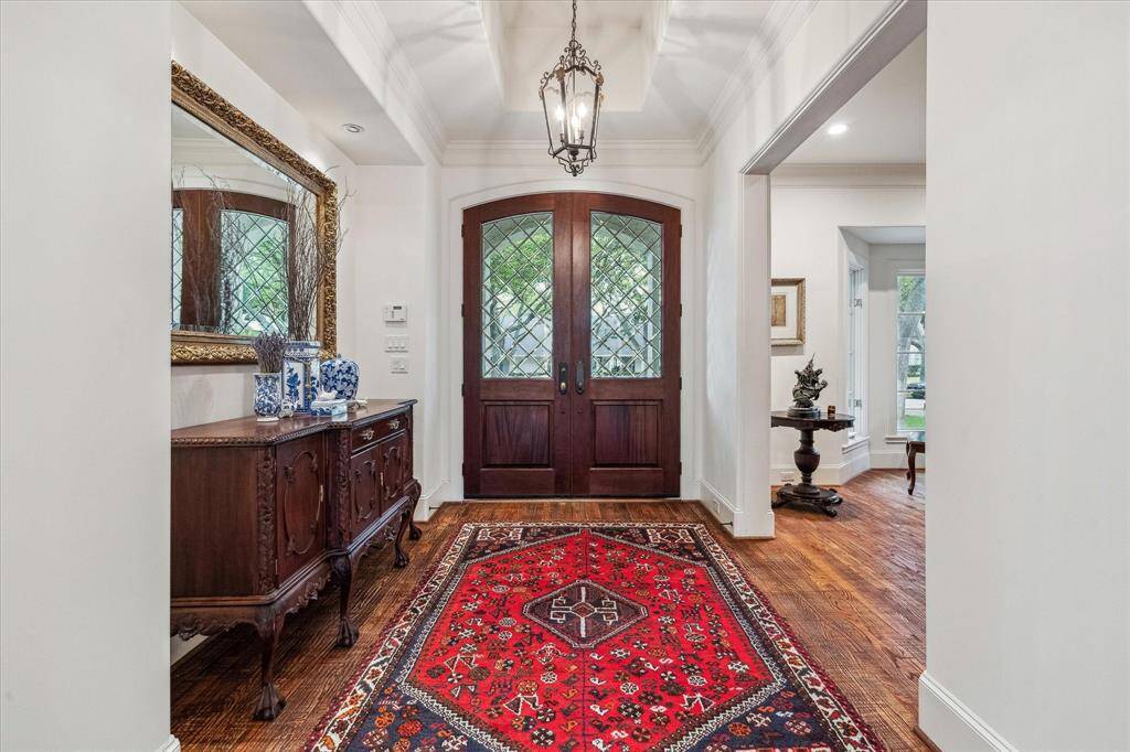 This elegant entryway features rich hardwood flooring, crisp white walls, and high ceilings with detailed crown molding. A coffered ceiling adds architectural interest, while a pair of wooden double doors with decorative glass panes welcomes natural light and creates a grand first impression.