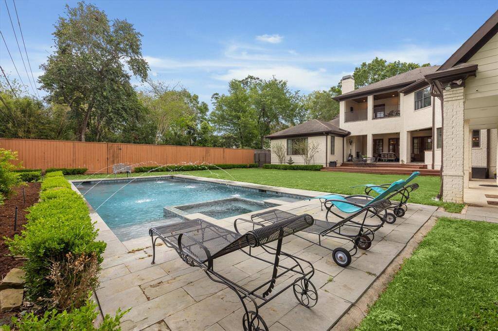 This pool and backyard area features a rectangular swimming pool with built-in fountains and an attached spa. It is surrounded by stone paving and a neatly landscaped lawn with trimmed hedges and mature trees. The space offers direct access to a spacious patio and a two-story covered outdoor living area.