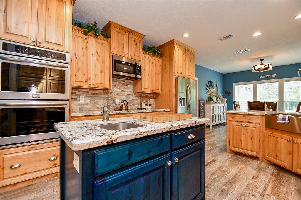 The island has a granite countertop with a built-in sink, and the space opens into a bright dining area with large windows and wood style tile flooring, creating a warm and inviting atmosphere.