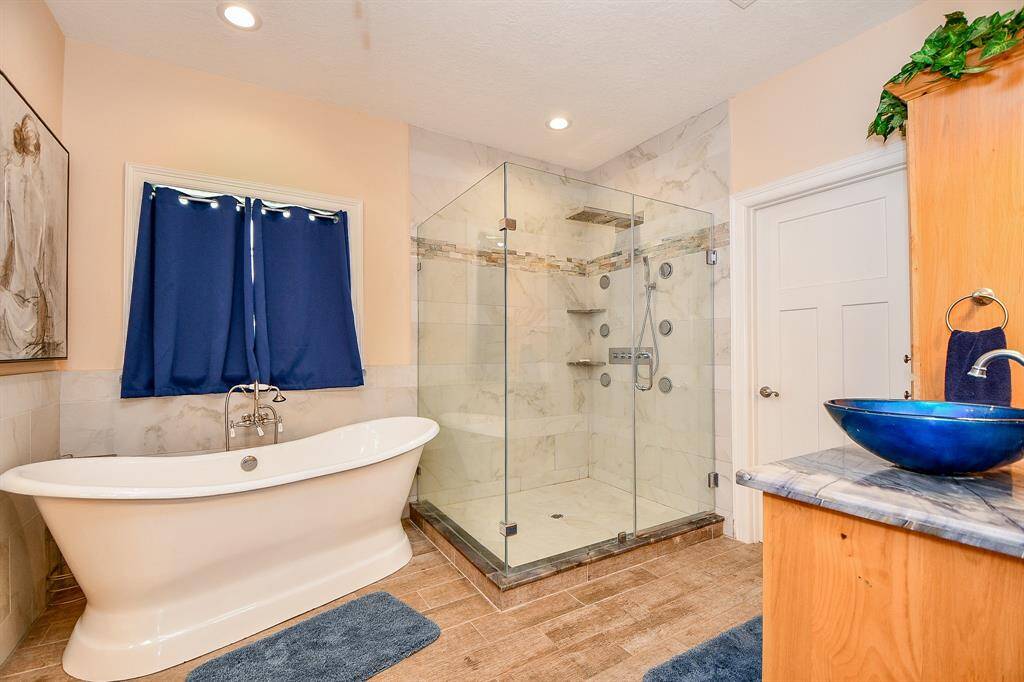 This modern bathroom features a sleek freestanding bathtub, a spacious glass-enclosed shower with multiple showerheads, and elegant tile work. A unique blue vessel sinks sit atop a wooden vanity, complementing the warm tones of the room.