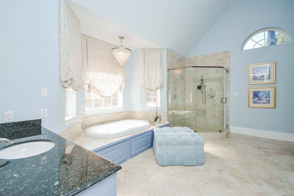 This Spacious Spa Like Bathroom features a luxurious soaking tub, High Ceilings, and Dual Vanities with Granite Countertops complemented by a stylish chandelier and ample natural light.