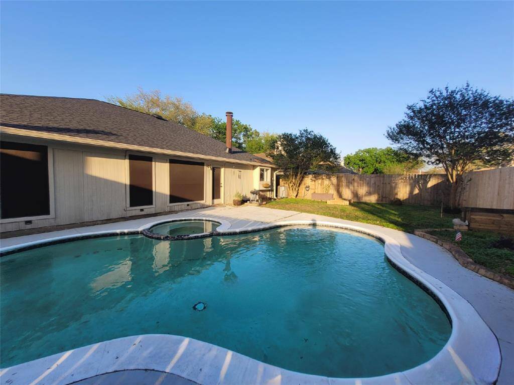 This backyard features a spacious pool with an attached spa, surrounded by a concrete deck. The area is fenced for privacy and includes grassy sections and mature trees, offering a blend of relaxation and outdoor enjoyment.