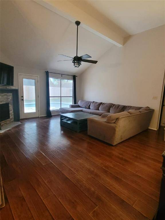 Spacious living room with high ceilings, wood flooring, and a cozy corner fireplace. Features a ceiling fan, and ample natural light from a glass door leading to the pool area.