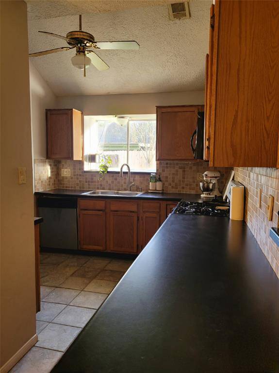 This kitchen features wooden cabinetry, a ceiling fan, and a large window above the sink for natural light. The space has tiled flooring and a sleek black countertop, providing a functional and inviting cooking area.