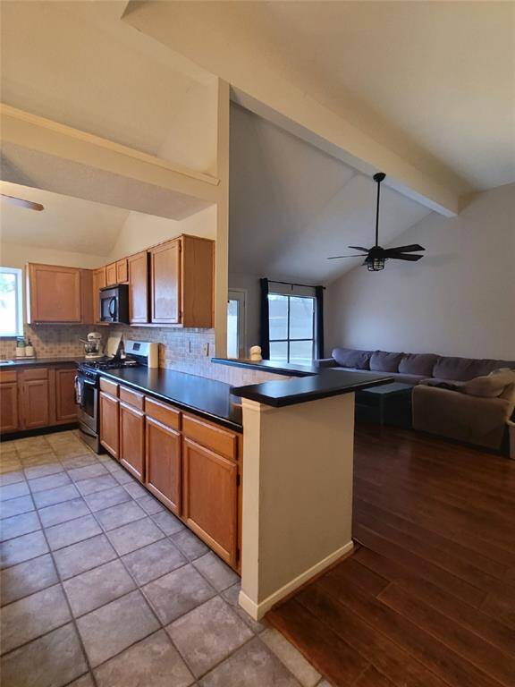 This photo shows a modern kitchen with wooden cabinets, tiled flooring, and a breakfast bar that opens to a cozy living area with hardwood floors and a ceiling fan. The space is bright and open, ideal for entertaining.