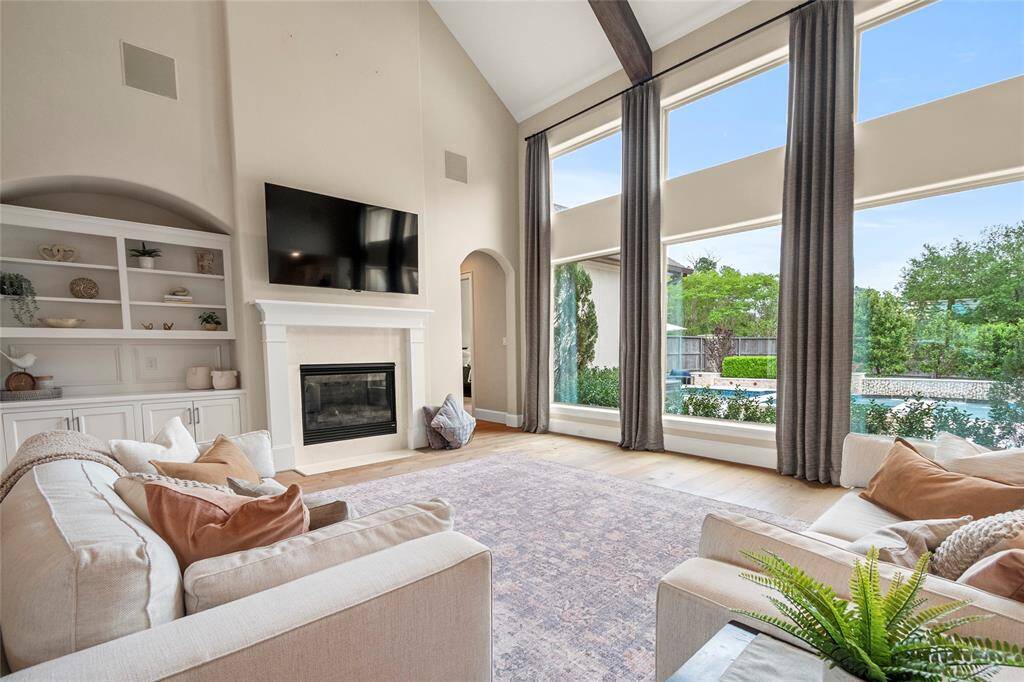 This spacious living room features high ceilings with exposed beams, a cozy fireplace, and built-in shelving. Large windows offer abundant natural light and a view of the pool and garden, creating a bright and inviting atmosphere.