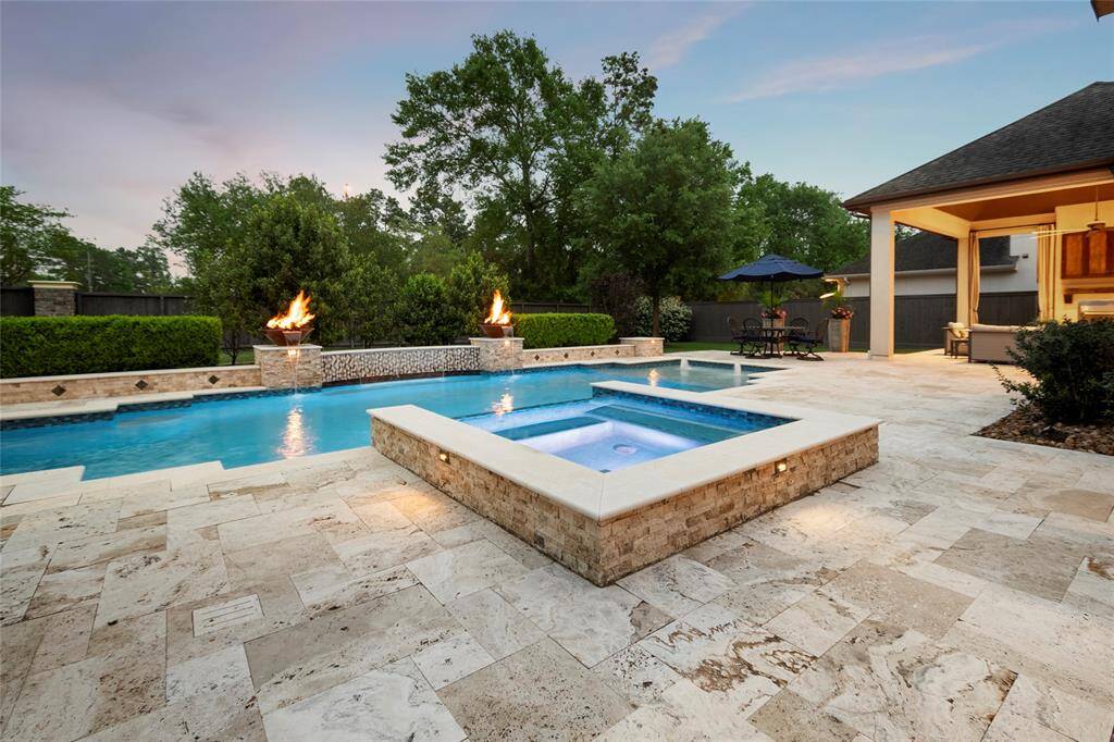 This photo showcases a luxurious backyard with a large swimming pool and spa, featuring elegant stone tile flooring. Two fire features add a touch of warmth and ambiance, while lush greenery and a covered patio with seating create an inviting outdoor entertainment space.