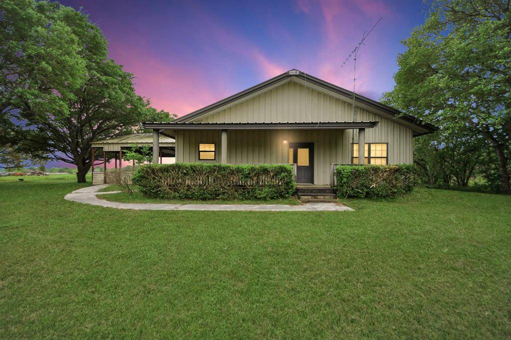 This photo shows a charming single-story home with a metal roof, nestled among lush greenery. The exterior features a welcoming front porch, neatly trimmed hedges, and a well-maintained lawn. The sky offers a beautiful sunset backdrop, enhancing the serene, countryside feel.