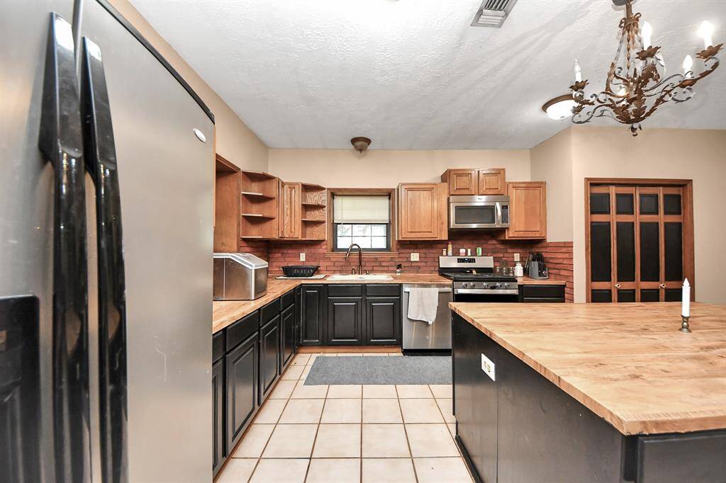 This kitchen features a spacious layout with a mix of black and wood cabinetry, stainless steel appliances, and a central island with butcher block countertops. The room is accented by a decorative chandelier and has a warm, inviting feel with tiled flooring.