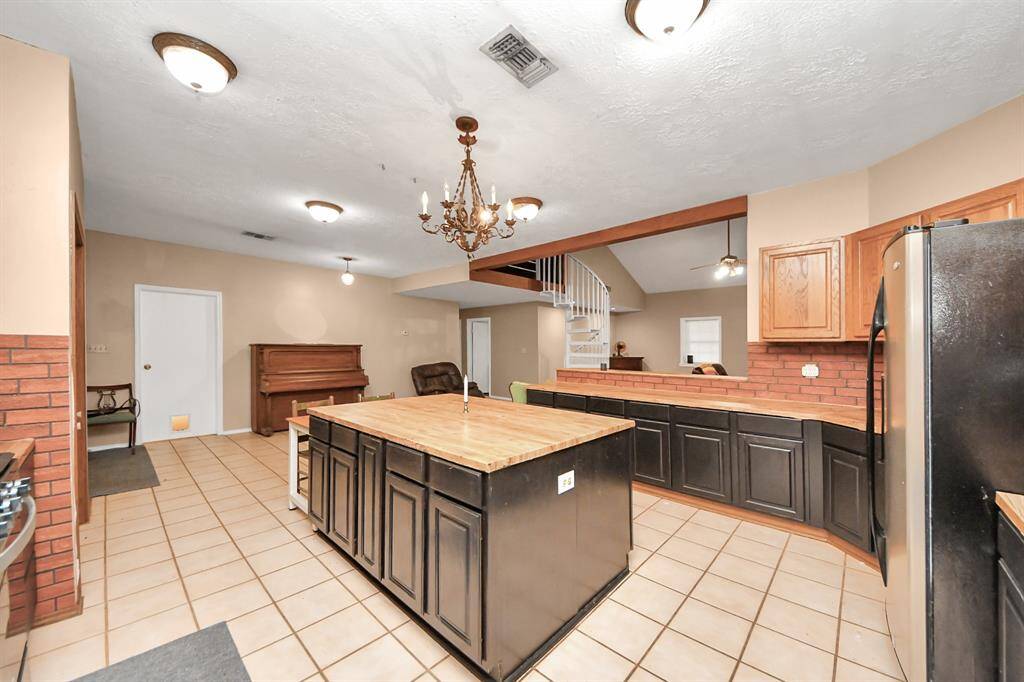 Spacious kitchen with a large central island, tile flooring, and dark cabinetry. Features a chandelier, stainless steel appliances, and a unique spiral staircase leading to an upper level. The open layout connects to a cozy living area.