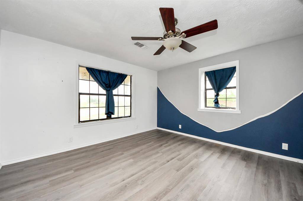This spacious primary bedroom features modern laminate flooring, a stylish ceiling fan, and two windows with blue curtains, allowing natural light to enhance its inviting atmosphere. The walls are a mix of white and blue, adding a touch of character.