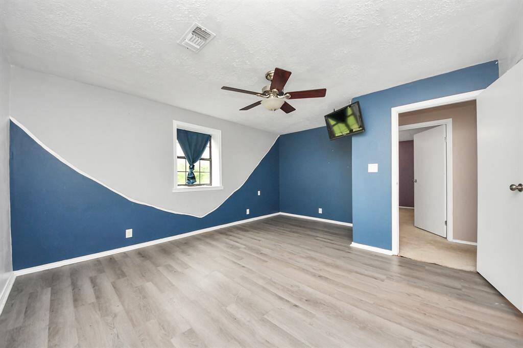 This bedroom features modern laminate flooring, a unique two-tone blue and white wall design, and a ceiling fan. A small window with a blue curtain provides natural light. The room connects to a carpeted hallway.