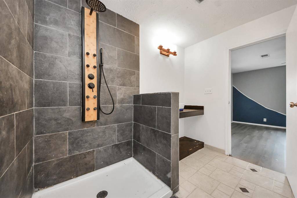 Modern bathroom with a sleek, dark-tiled shower featuring a rainfall showerhead. Adjacent is a small vanity area, and the room opens to a space with light-colored flooring and a creatively painted wall.
