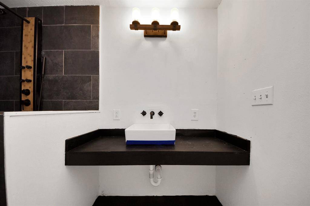 This bathroom features a modern black countertop with a white vessel sink, complemented by a sleek wall-mounted faucet. The shower area is tiled in dark tones, adding a contemporary touch. A stylish light fixture above enhances the space.