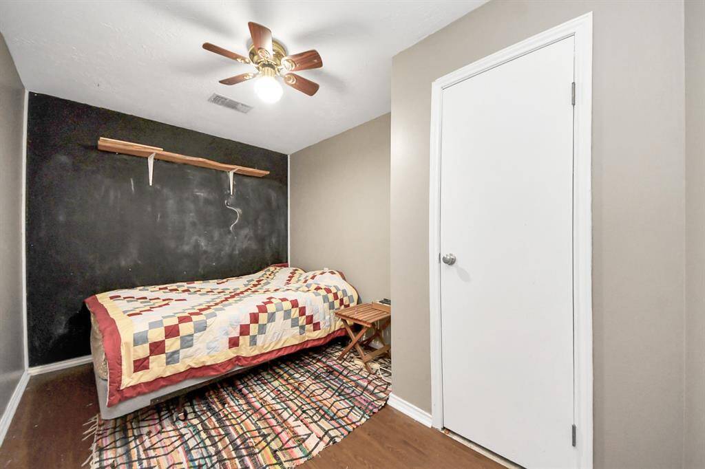 Cozy bedroom featuring a black accent wall with a wooden shelf, a ceiling fan, and hardwood floors. A bed with a colorful quilt is placed on a patterned rug, creating a warm and inviting atmosphere.
