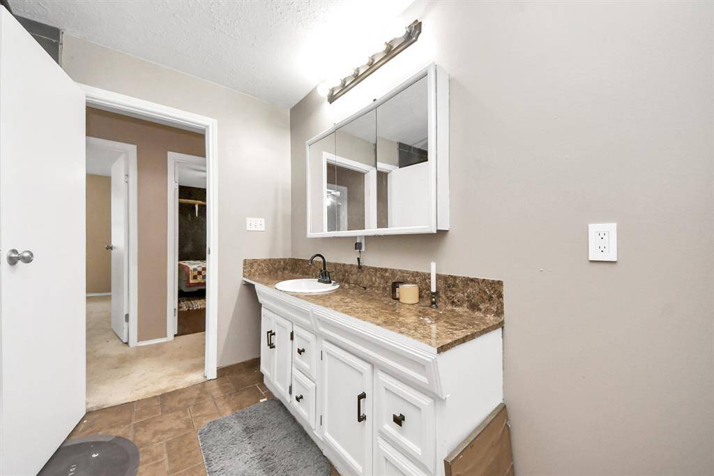 This bathroom features a spacious vanity with a brown countertop and white cabinetry, complemented by a large mirror and overhead lighting. The neutral wall color and tile flooring create a clean, modern look.