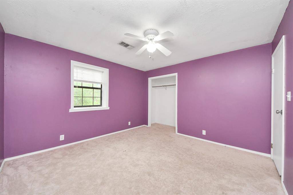 Bright bedroom with purple walls, soft carpet, and a ceiling fan. Features include a window with natural light and a small closet.