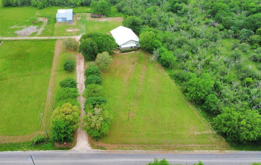 This aerial photo shows a spacious rural property surrounded by lush greenery and trees. A long driveway leads from the main road to the building, with open grassy areas and additional structures nearby, offering privacy and ample space.