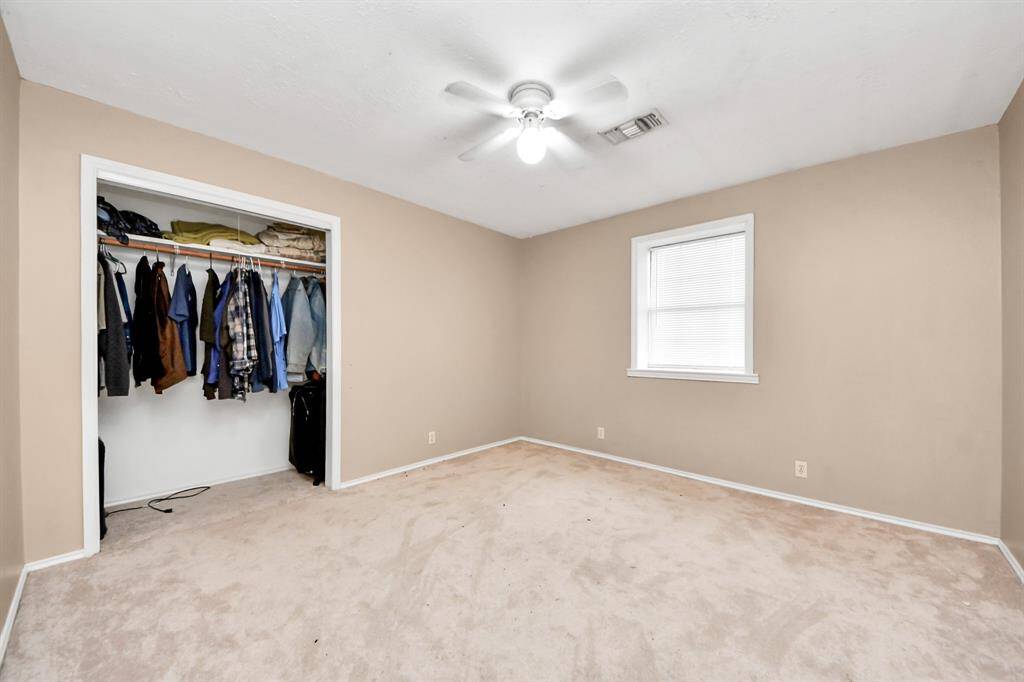 This photo shows a cozy bedroom with neutral-colored walls and carpet. It features a ceiling fan and a window for natural light. There is an open closet with ample hanging space.