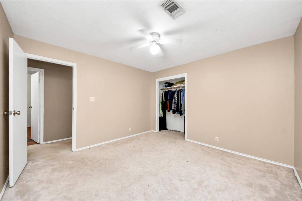 This photo shows a simple, carpeted bedroom with neutral walls, a ceiling fan, and an open closet. It has a door leading to another room or hallway.