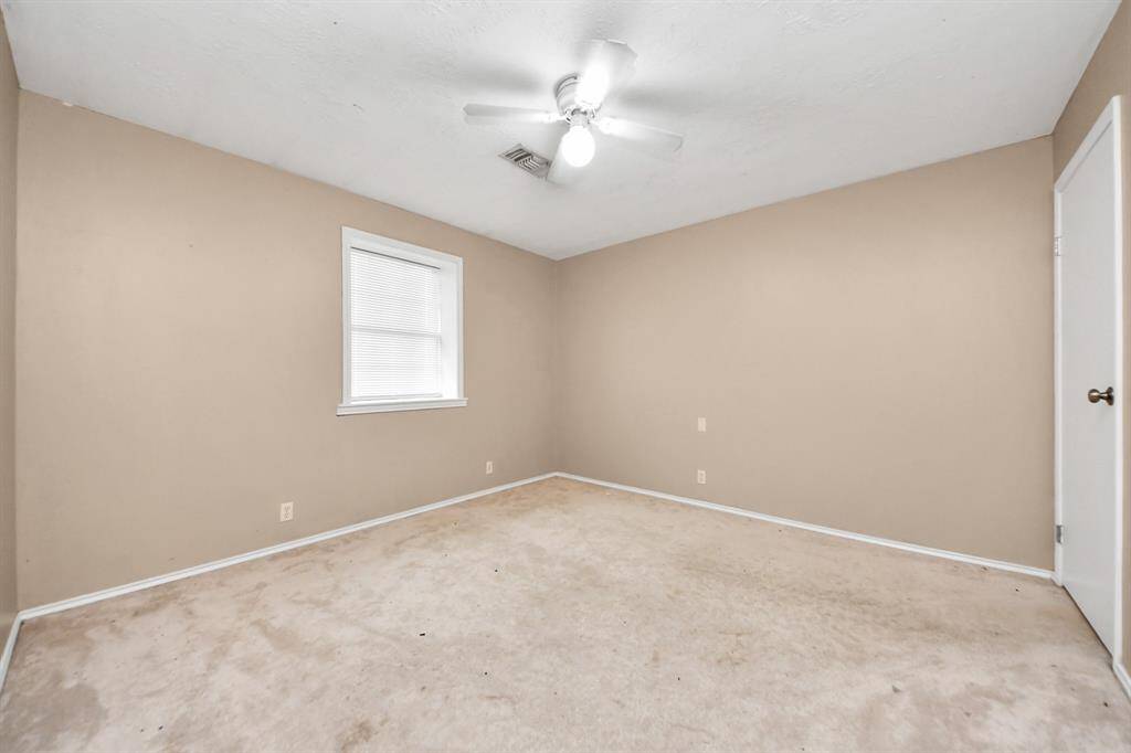 This is a basic, empty room with neutral beige walls and carpet, featuring a white ceiling fan and a window with blinds. Ideal for customization.