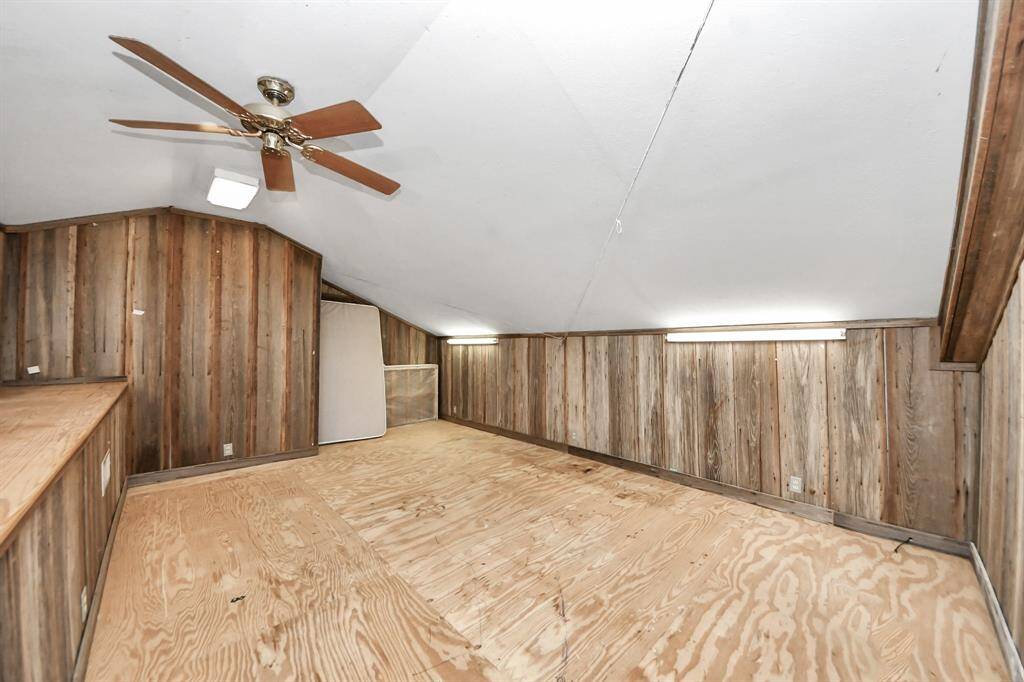 This photo shows a cozy attic space with wood panel walls and a sloped ceiling. It features a ceiling fan, bright lighting, and a simple plywood floor, offering potential for customization as a storage area or cozy retreat.